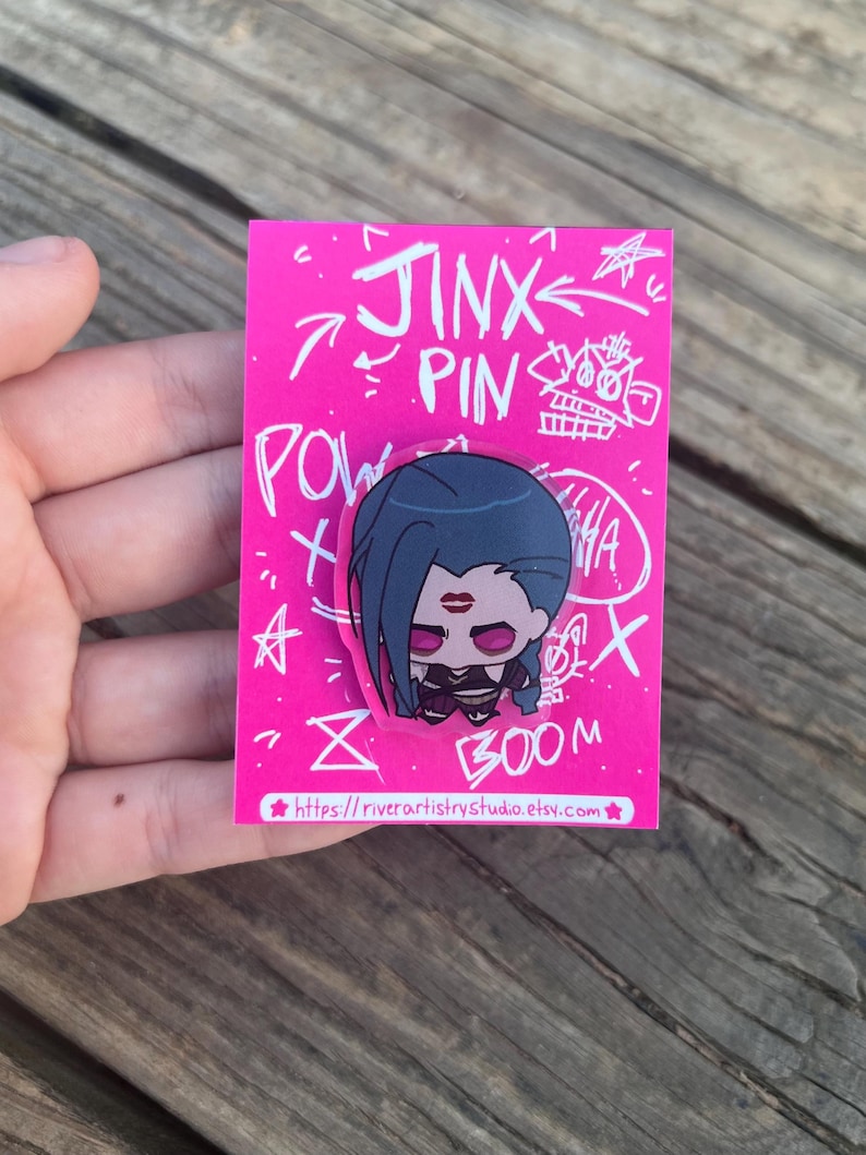Pode incluir: Um alfinete rosa com um personagem de desenho animado com cabelo azul, batom vermelho e uma roupa preta. O alfinete est&aacute; em um cart&atilde;o rosa com as palavras "Jinx Pin" e outros rabiscos. O endere&ccedil;o do site &eacute; vis&iacute;vel na parte inferior.