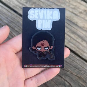 Pode incluir: Um pin preto com uma ilustra&ccedil;&atilde;o de desenho animado de um personagem com cabelo escuro e batom vermelho. O pin est&aacute; em um cart&atilde;o preto com o texto "SEVIKA PIN" em branco e um endere&ccedil;o de site na parte inferior.