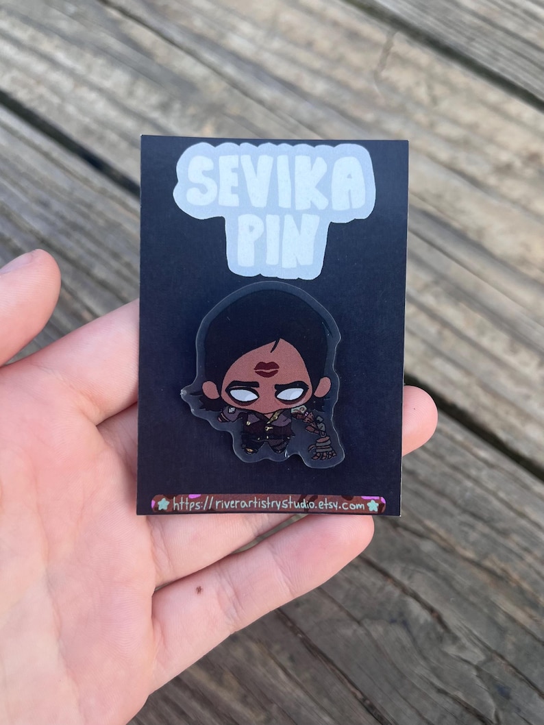 Pode incluir: Um pin preto com uma ilustra&ccedil;&atilde;o de desenho animado de um personagem com cabelo escuro e batom vermelho. O pin est&aacute; em um cart&atilde;o preto com o texto "SEVIKA PIN" em branco e um endere&ccedil;o de site na parte inferior.