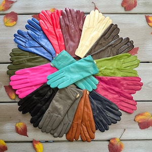 Peut inclure: Une présentation circulaire de gants en cuir de différentes couleurs : rouge, bleu, vert et noir. Les gants sont disposés sur une surface en bois avec des feuilles d'automne éparpillées, mettant en valeur la diversité des couleurs.