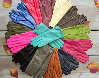 Peut inclure: Une présentation circulaire de gants en cuir de différentes couleurs : rouge, bleu, vert et noir. Les gants sont disposés sur une surface en bois avec des feuilles d'automne éparpillées, mettant en valeur la diversité des couleurs.