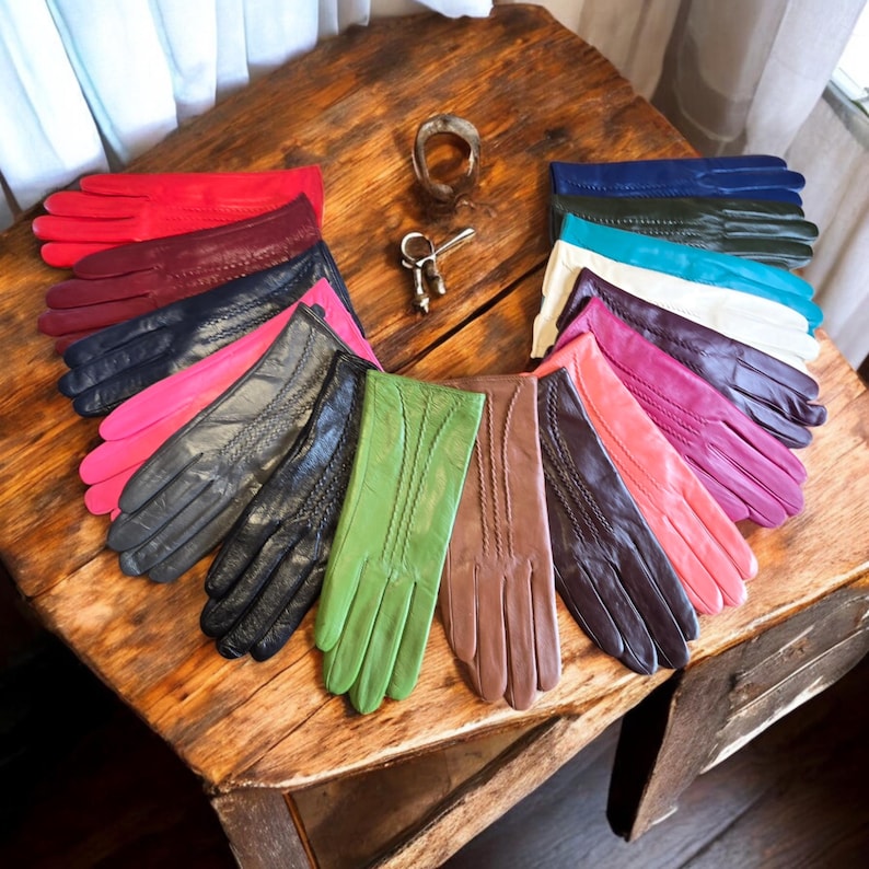 May include: A collection of 12 pairs of leather gloves in various colors, including red, pink, teal, white, purple, brown, green, and black. The gloves are arranged in a semi-circle on a wooden surface.