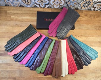 Peut inclure: Une variété de gants en cuir de différentes couleurs, notamment noir, marron, rose, rouge, vert et bleu. Les gants sont disposés en éventail sur une surface en bois. Le texte "TheGlov" est visible sur une boîte en arrière-plan.