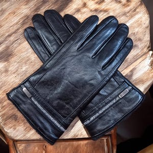 Könnte beinhalten: Ein Paar schwarze Lederhandschuhe mit weißem und grauem Besatz. Die Handschuhe haben einen Reißverschluss-Detail am Handgelenk.