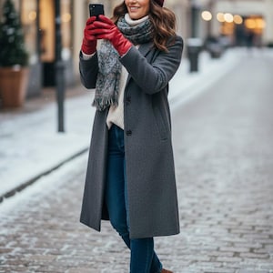 Könnte beinhalten: Eine Person mit einer weinroten Strickmütze, roten Handschuhen und einem grauen Mantel, die ein Smartphone hält. Sie trägt außerdem einen weißen Pullover, einen gemusterten Schal, blaue Jeans und braune Stiefel. Die Szene spielt auf einer Kopfsteinpflasterstraße.
