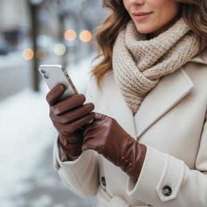 May include: A person wearing brown leather gloves, a beige scarf, and a cream-coloured coat. The gloves are buttoned at the wrist. They are holding a silver smartphone. The background is blurred with a snowy, outdoor scene.