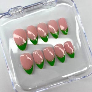 May include: A set of ten false nails in a clear plastic case. The nails are almond shaped and have a light pink base with a green tip and a thin gold glitter line.