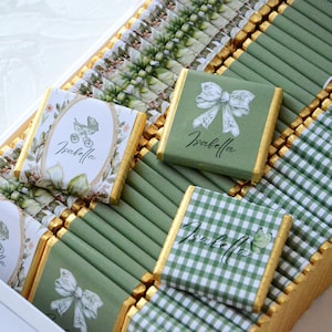 May include: A collection of square chocolate bars wrapped in sage green and white paper. Designs include a baby carriage, a decorative bow, and a green gingham pattern. Each wrapper features the name Isabella in a cursive black script font.