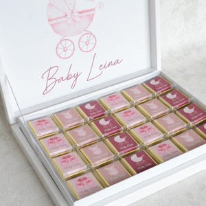 May include: A white box containing chocolates, ideal for a baby celebration. The lid displays a pink baby carriage illustration and the name "Baby Leina". Individual chocolates are decorated with baby-related images and text.