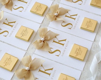May include: Arrangement of white cards with gold lettering and small gold-wrapped chocolates. The cards feature the initials "SK" and the names "SARAH & KEVIN". Each card is adorned with a gold ribbon bow and a pearl.