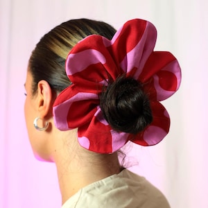 May include: A red and pink patterned fabric scrunchie in the shape of a flower. The scrunchie is worn in a woman's hair.