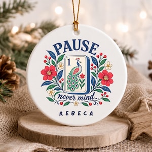 May include: A round white ceramic ornament featuring a central illustration of a mahjong tile with a peacock and the number one. The design includes red flowers, green leaves, and the text PAUSE never mind... REBECA in dark blue lettering.