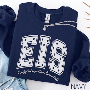 May include: A navy blue crewneck sweatshirt featuring the large white letters EIS with a polka dot pattern. Below the letters, the text Early Intervention Specialist is printed in a white script font. A silver chain necklace rests on the collar.