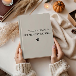 May include: A beige "Pawprints on My Heart" pet memory journal. The journal has the text "PET MEMORY JOURNAL" and a paw print graphic. The journal is held by a person wearing a cream-colored sweater. The background includes candles and a small pumpkin.