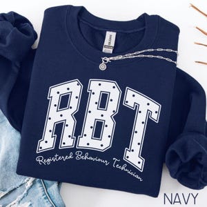 May include: A navy blue crewneck sweatshirt featuring the white text RBT in a large collegiate font with a polka dot pattern inside the letters. Below the letters, the text Registered Behaviour Technician is written in a white script font.