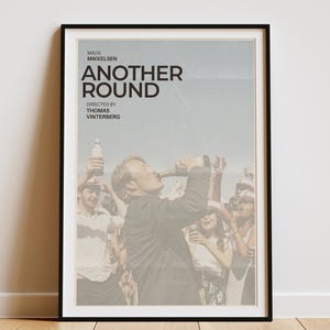May include: A framed poster for the film "Another Round" featuring Mads Mikkelsen. The poster shows a man drinking from a bottle, surrounded by a crowd. The film title and credits are displayed at the top.