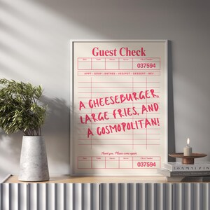 May include: A framed print resembling a restaurant guest check, featuring the text "Guest Check" at the top and a list of food items: "A CHEESEBURGER, LARGE FRIES, AND A COSMOPOLITAN!" The print is displayed on a wooden surface.
