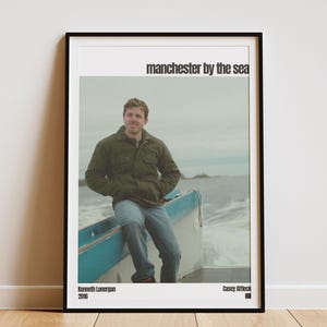 May include: A framed poster featuring a man sitting in a boat with the text "manchester by the sea". The poster has a black frame and a white border. The poster also includes the names Kenneth Lonergan and Casey Affleck, and the year 2016.