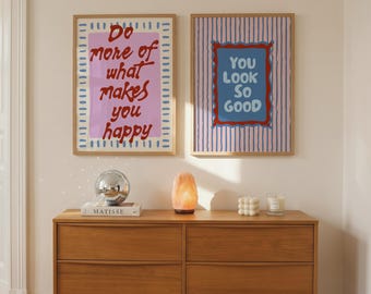 May include: Two framed rectangular wall art prints with light wood frames. The left print has a pink background with blue dashed borders and red text reading Do more of what makes you happy. The right print has blue stripes and blue text reading YOU LOOK SO GOOD.
