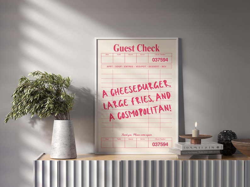 May include: A framed print resembling a restaurant guest check, featuring the text "Guest Check" at the top and a list of food items: "A CHEESEBURGER, LARGE FRIES, AND A COSMOPOLITAN!" The print is displayed on a wooden surface.