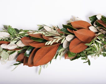Op de afbeelding: Een krans van magnolia bladeren, olijftakken en eucalyptus takjes. De bladeren zijn donkerbruin, de olijftakken zijn groen en de eucalyptus takjes zijn lichtgrijs.
