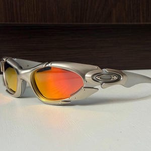 Puede incluir: Gafas de sol plateadas con lentes naranjas y amarillas. Las gafas de sol tienen un dise&ntilde;o futurista con una montura curvada y una est&eacute;tica elegante y moderna. Las lentes reflejan un degradado de colores.
