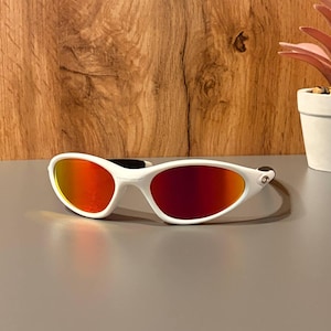 Könnte beinhalten: Weiße Sonnenbrille mit sportlichem Design und leuchtend rot-orangen Gläsern. Die Sonnenbrille hat einen geschwungenen Rahmen und einen schwarzen Steg. Das Bild ist vor einem Holz- und einem grauen Hintergrund platziert.