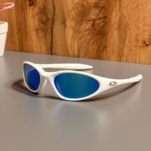 Peut inclure: Lunettes de soleil blanches avec verres bleus. Les lunettes ont un design moderne avec une monture incurvée et un petit logo sur les branches. Les verres sont d'un bleu vif et la monture est blanche.