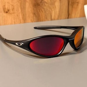 Puede incluir: Gafas de sol negras con lentes rojas y naranjas. Las gafas de sol tienen un diseño elegante y moderno con una montura negra y patillas curvas. Las lentes hacen una transición de rojo a naranja, ofreciendo un aspecto elegante.