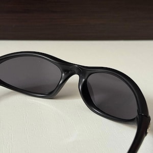 Puede incluir: Gafas de sol negras con lentes de color gris oscuro. Las gafas tienen una montura curvada y un dise&ntilde;o elegante y moderno. Las lentes son de forma ovalada y parecen tintadas. Las gafas probablemente son de pl&aacute;stico.