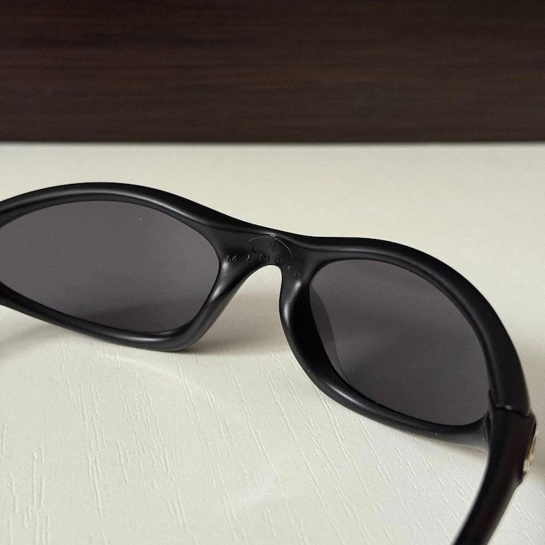 Puede incluir: Gafas de sol negras con lentes de color gris oscuro. Las gafas tienen una montura curvada y un dise&ntilde;o elegante y moderno. Las lentes son de forma ovalada y parecen tintadas. Las gafas probablemente son de pl&aacute;stico.
