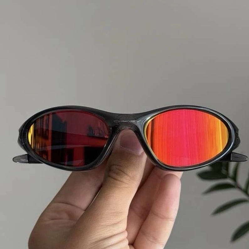 Puede incluir: Gafas de sol ovaladas negras con lentes rojas. Las gafas tienen una montura oscura y se sostienen en una mano. Las lentes reflejan el entorno. Un accesorio de moda.