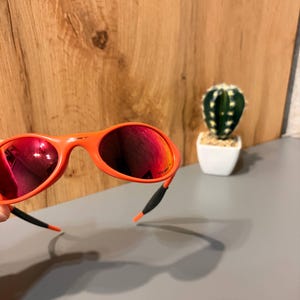 May include: Orange sunglasses with red mirrored lenses. The sunglasses have a curved frame and black and orange ear pieces. A small cactus in a white pot is in the background.