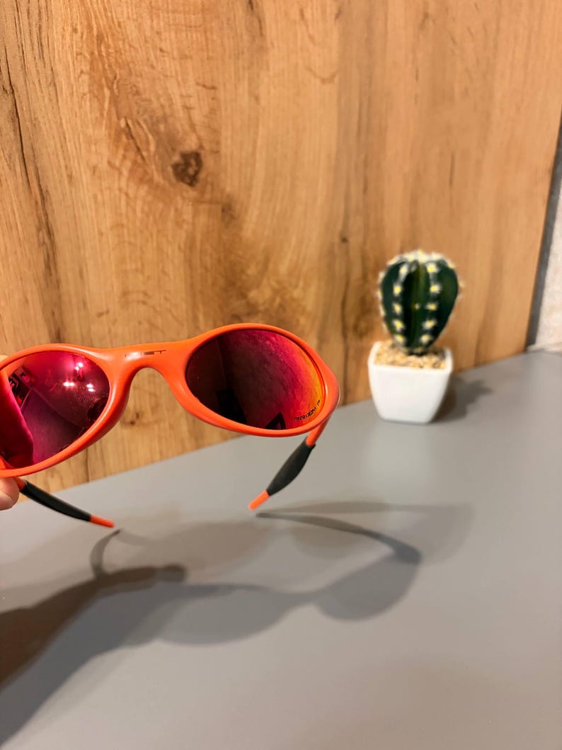May include: Orange sunglasses with red mirrored lenses. The sunglasses have a curved frame and black and orange ear pieces. A small cactus in a white pot is in the background.