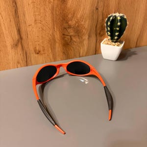 May include: Orange sunglasses with black lenses and black temple tips. The sunglasses are set against a gray surface with a wooden background and a small cactus in a white pot.