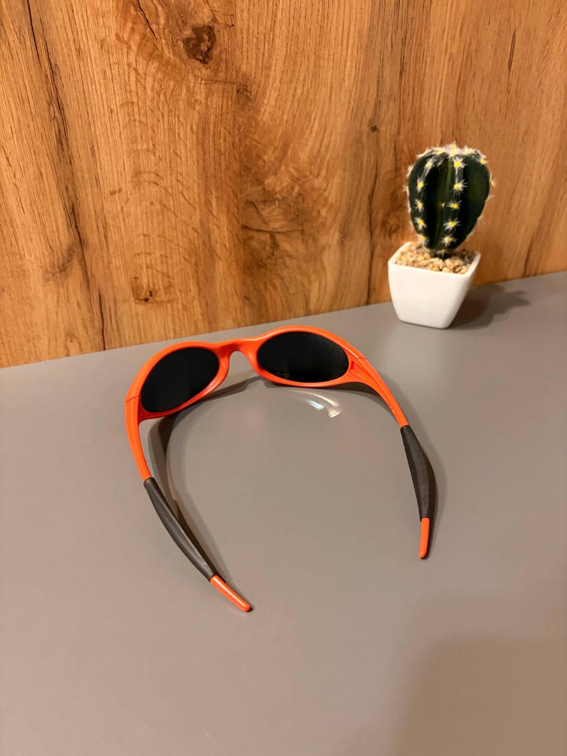 May include: Orange sunglasses with black lenses and black temple tips. The sunglasses are set against a gray surface with a wooden background and a small cactus in a white pot.