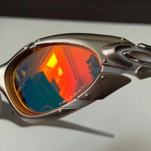 Puede incluir: Primer plano de gafas de sol plateadas con lentes polarizadas tintadas en rojo. Las lentes reflejan una luz naranja y amarilla brillante. La montura tiene un acabado met&aacute;lico con remaches y un dise&ntilde;o curvo.