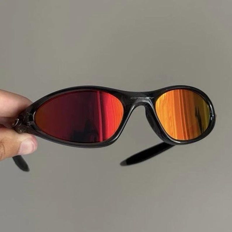 Puede incluir: Gafas de sol negras con lentes degradadas rojas y naranjas. Las gafas tienen un dise&ntilde;o elegante y deportivo, con una montura curvada y un peque&ntilde;o logotipo en la barra superior. Las lentes hacen la transici&oacute;n del rojo al naranja.