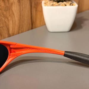May include: Close-up of orange sunglasses with black earpieces. The text "CE MADE IN U.S.A." is printed on the arm of the sunglasses. The sunglasses are set against a neutral background.