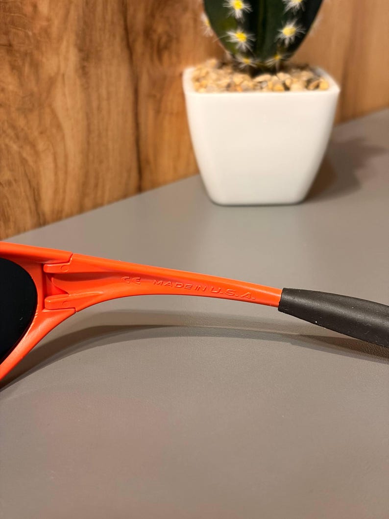 May include: Close-up of orange sunglasses with black earpieces. The text "CE MADE IN U.S.A." is printed on the arm of the sunglasses. The sunglasses are set against a neutral background.