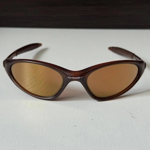 May include: Brown sunglasses with a curved frame and tinted lenses. The brand name "OAKLEY" is printed on the frame. The sunglasses are designed for eye protection and fashion.