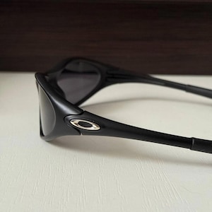 Puede incluir: Gafas de sol negras con acabado mate. Las gafas tienen un dise&ntilde;o curvado con lentes oscuras y un emblema plateado en la montura. Las patillas de las gafas son finas y se extienden hacia atr&aacute;s.