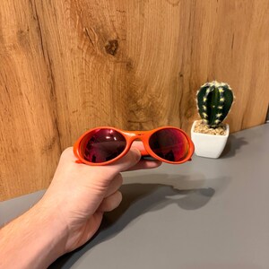 May include: Orange sunglasses with red-tinted lenses. The sunglasses are held in a hand. A small cactus in a white pot is in the background. The sunglasses have a rounded frame.