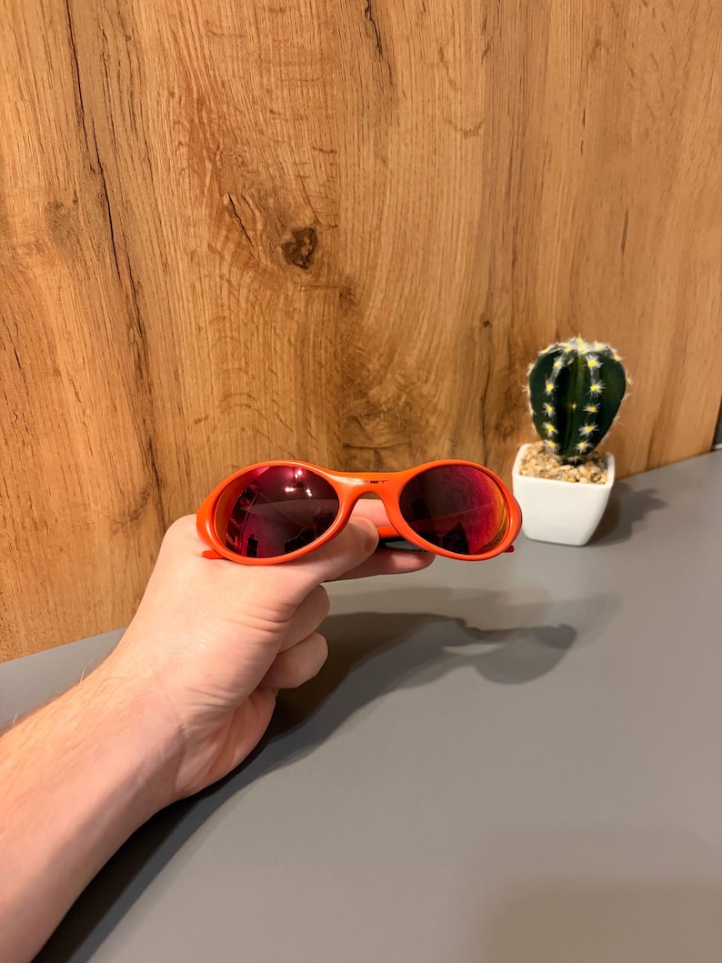 May include: Orange sunglasses with red-tinted lenses. The sunglasses are held in a hand. A small cactus in a white pot is in the background. The sunglasses have a rounded frame.