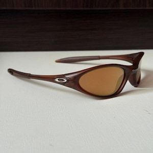 May include: Brown sunglasses with a curved frame and tinted lenses. The sunglasses have a sleek, modern design with a small silver emblem on the side of the frame. The lenses are a warm, amber color.