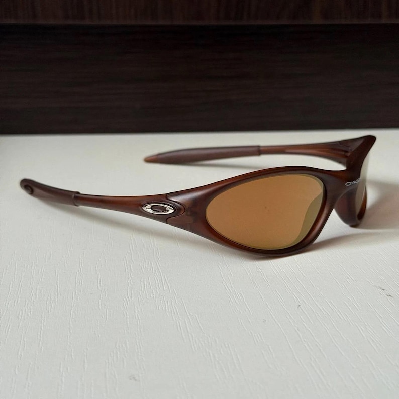 May include: Brown sunglasses with a curved frame and tinted lenses. The sunglasses have a sleek, modern design with a small silver emblem on the side of the frame. The lenses are a warm, amber color.