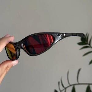 Puede incluir: Gafas de sol negras y rojas con montura curvada. Las lentes son de color rojo vibrante y la montura tiene un dise&ntilde;o elegante y moderno. Las gafas de sol est&aacute;n sujetas por una mano.