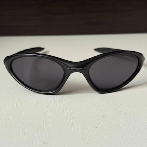 Puede incluir: Gafas de sol negras con montura curvada y lentes oscuras. El nombre de la marca "Oakley" se muestra sutilmente en el puente. Las gafas de sol tienen un dise&ntilde;o elegante y moderno.