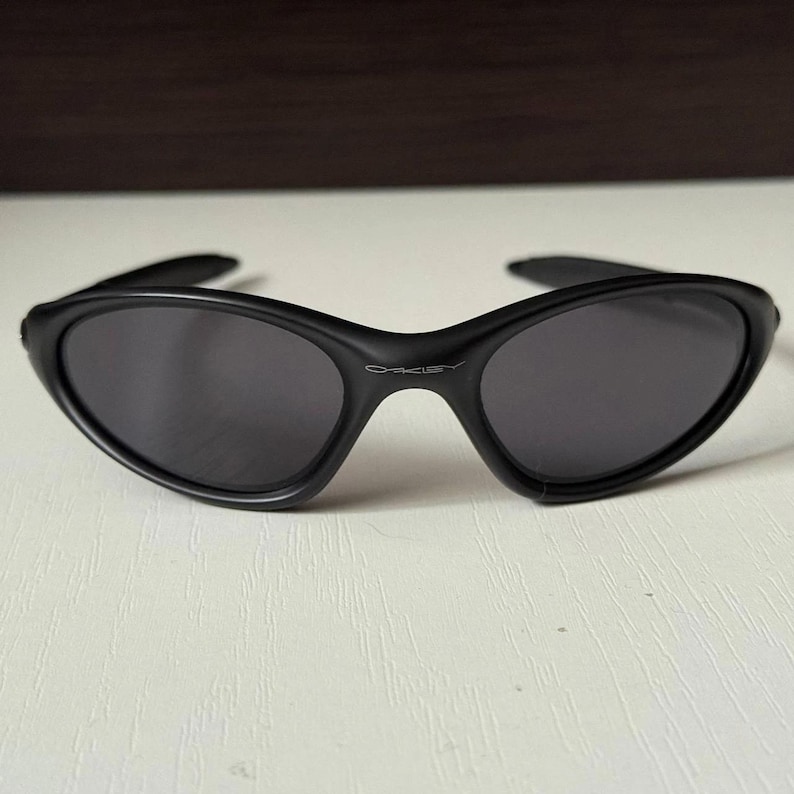 Puede incluir: Gafas de sol negras con montura curvada y lentes oscuras. El nombre de la marca "Oakley" se muestra sutilmente en el puente. Las gafas de sol tienen un dise&ntilde;o elegante y moderno.