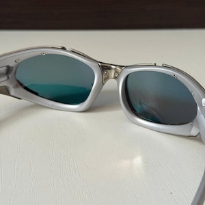 Puede incluir: Gafas de sol plateadas con lentes azul-verdosas. Las gafas de sol tienen un dise&ntilde;o elegante y moderno con un acabado met&aacute;lico. Las lentes son reflectantes y la montura tiene una forma curvada.